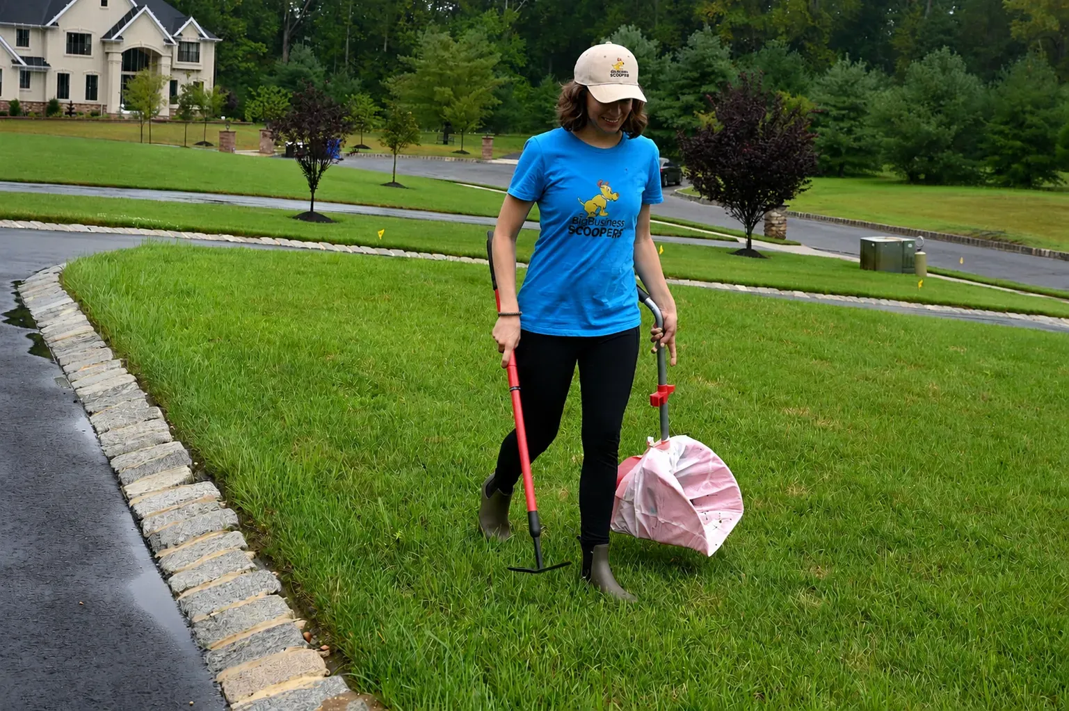 Owner Emily LaBeaume in a branded blue shirt raking up dog waste on a freshly mowed New Jersey lawn